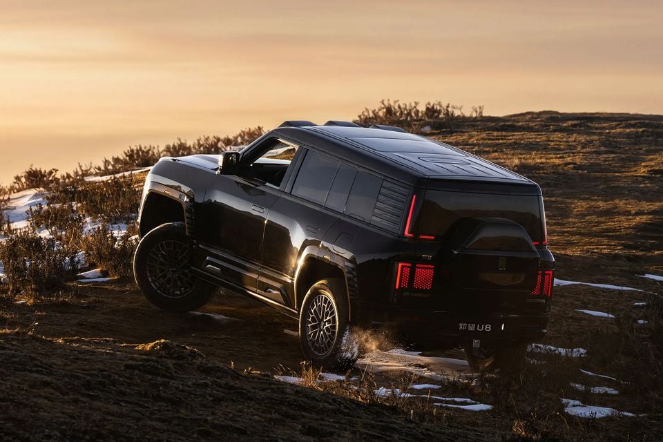 A black Yangwang U8 SUV driving up a snowy, grassy hillside at sunset, seen from the rear quarter